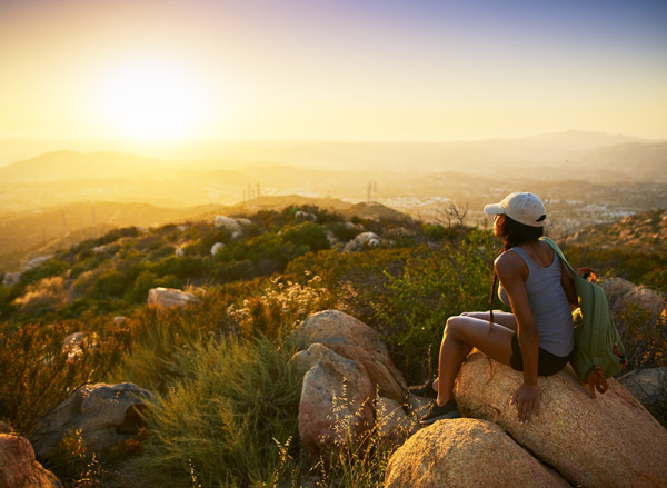Photo of rear view of woman hiker sitting on rock on top of hill while looking at sunset over San Diego California